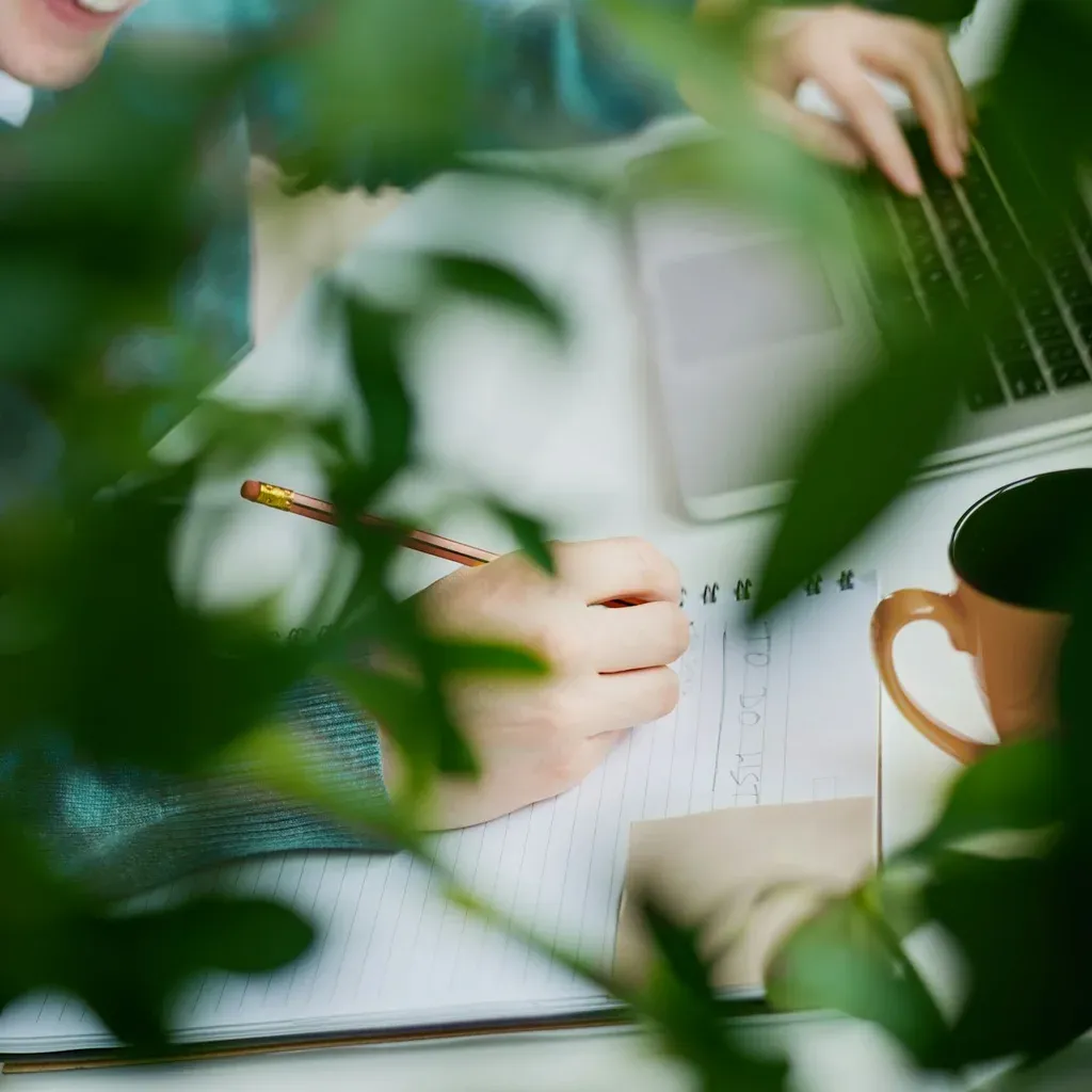 Someone writing notes by hand beside a laptop, seen through leaves