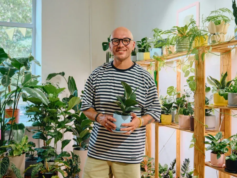 A shop owner standing in his plant shop holding a potted plant, shelves of plants behind him