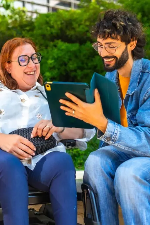 Two people sitting outdoors looking at a tablet together, one of them using a power wheelchair